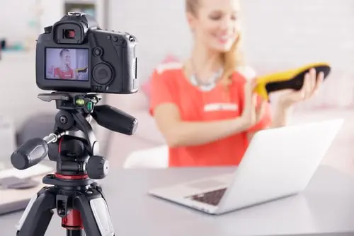 A woman recording a video with a DSLR camera on a tripod, showcasing a product while sitting at a desk with a laptop. The image represents content creation, video marketing, and influencer product reviews.